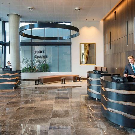 Pullman Brisbane Airport Hotell Exteriör bild The image shows a modern hotel lobby. There are two reception desks made of dark-colored materials, and staff members are attending to guests at these desks. The lobby features large windows that allow natural light to fill the space, and there are p