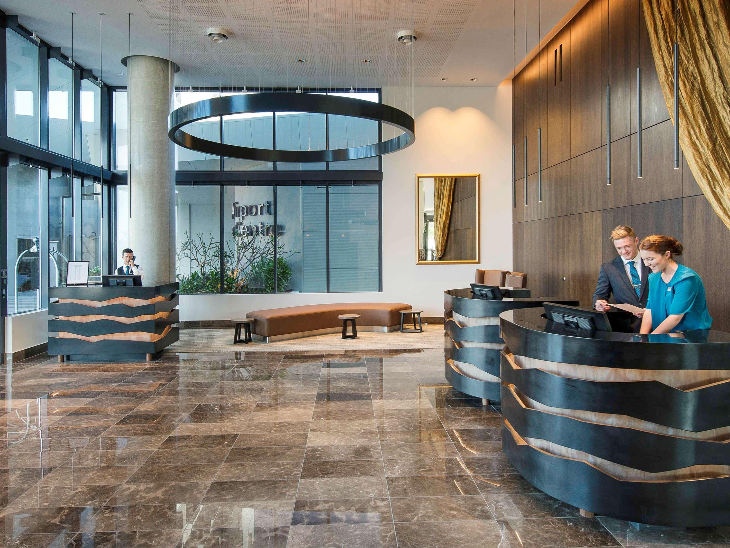 Pullman Brisbane Airport Hotell Exteriör bild The image shows a modern hotel lobby. There are two reception desks made of dark-colored materials, and staff members are attending to guests at these desks. The lobby features large windows that allow natural light to fill the space, and there are p